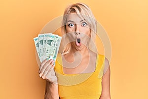 Young blonde girl holding 10000 south korean won banknotes scared and amazed with open mouth for surprise, disbelief face