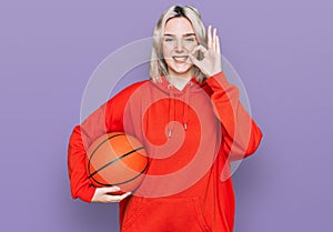 Young blonde girl holding basketball ball doing ok sign with fingers, smiling friendly gesturing excellent symbol