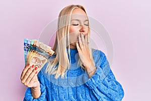 Young blonde girl holding canadian dollars bored yawning tired covering mouth with hand