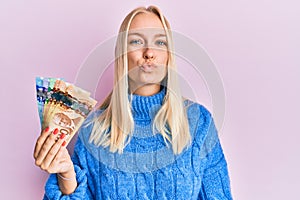 Young blonde girl holding canadian dollars looking at the camera blowing a kiss on air being lovely and sexy