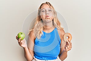 Young blonde girl holding green apple and donut puffing cheeks with funny face