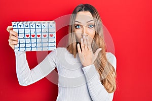 Young blonde girl holding heart calendar covering mouth with hand, shocked and afraid for mistake