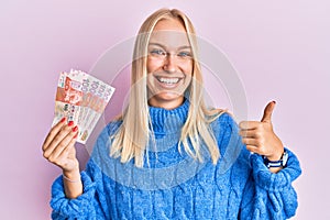 Young blonde girl holding hong kong 10 dollars banknotes smiling happy and positive, thumb up doing excellent and approval sign