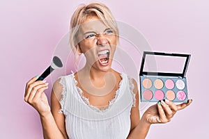 Young blonde girl holding makeup brush and blush angry and mad screaming frustrated and furious, shouting with anger