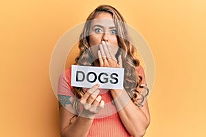 Young blonde girl holding paper with dogs message covering mouth with hand, shocked and afraid for mistake