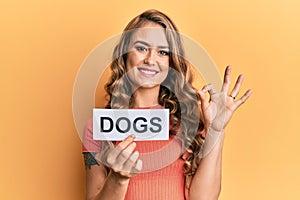 Young blonde girl holding paper with dogs message doing ok sign with fingers, smiling friendly gesturing excellent symbol