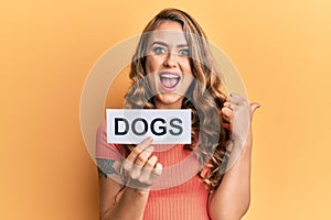 Young blonde girl holding paper with dogs message pointing thumb up to the side smiling happy with open mouth