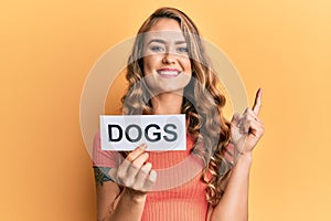 Young blonde girl holding paper with dogs message smiling with an idea or question pointing finger with happy face, number one