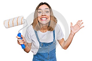 Young blonde girl holding roller painter celebrating victory with happy smile and winner expression with raised hands