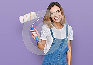 Young blonde girl holding roller painter looking positive and happy standing and smiling with a confident smile showing teeth