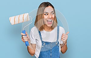 Young blonde girl holding roller painter pointing thumb up to the side smiling happy with open mouth