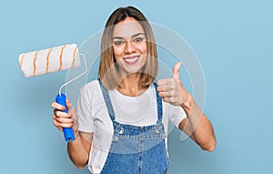 Young blonde girl holding roller painter smiling happy and positive, thumb up doing excellent and approval sign