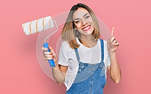 Young blonde girl holding roller painter surprised with an idea or question pointing finger with happy face, number one