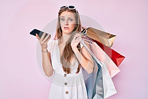 Young blonde girl holding shopping bags talking on the smartphone clueless and confused expression