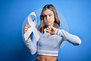 Young blonde girl holding stop women violence and lung cancer awareness white ribbon with angry face, negative sign showing