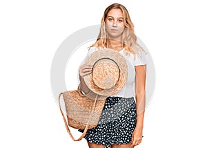 Young blonde girl holding summer hat and wicker handbag thinking attitude and sober expression looking self confident