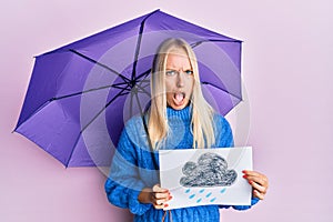 Young blonde girl holding umbrella rain draw in shock face, looking skeptical and sarcastic, surprised with open mouth