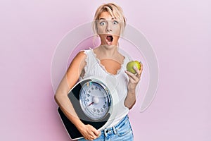 Young blonde girl holding weight machine and green apple afraid and shocked with surprise and amazed expression, fear and excited