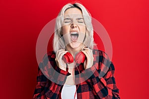 Young blonde girl listening to music using headphones angry and mad screaming frustrated and furious, shouting with anger looking