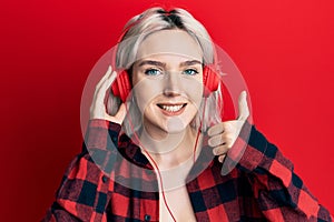 Young blonde girl listening to music using headphones smiling happy and positive, thumb up doing excellent and approval sign