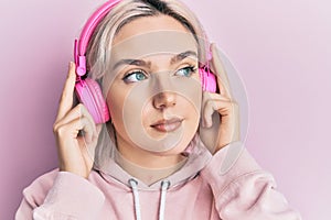 Young blonde girl listening to music using headphones smiling looking to the side and staring away thinking