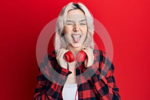 Young blonde girl listening to music using headphones sticking tongue out happy with funny expression