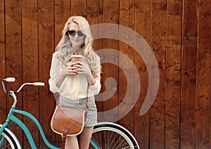 Young blonde girl with long hair with brown vintage bag in sunglasses standing near vintage green bicycle and holding a cup o