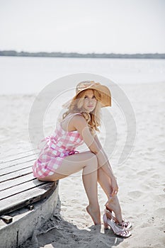 Young blonde girl look like a Barbie doll in pink mini dress and wide brim hat sitting barefoot on beach