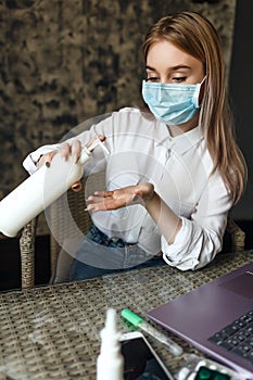 A young blonde girl in a medical mask sits in a room at a table with a laptop, medicine and uses an antiseptic