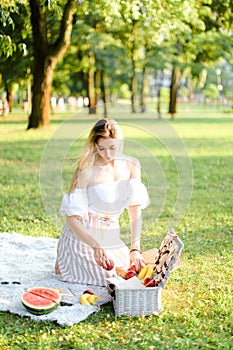 Young blonde girl preparing forpicnic in park, sitting on plaid near box and waterlemon.