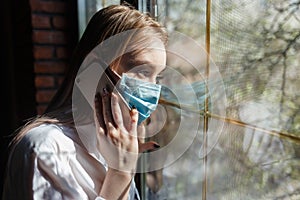 Young blonde girl in a protective mask in a dark room looks out the window. Self-isolation from the coronovirus pandemic