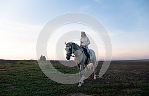 Young blonde girl riding on a horse on the field during sunset