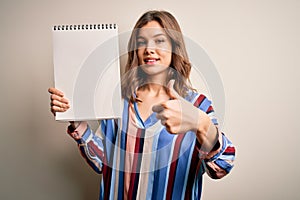 Young blonde girl showing white space paper on notebook over isolated background happy with big smile doing ok sign, thumb up with