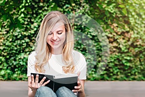 Young blonde student girl sits on a bench, reads a book and smiles in a park