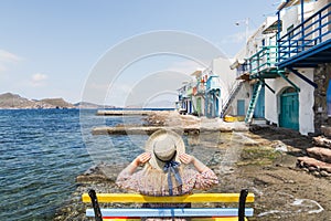 Young blonde girl sitting on a bench overlooking colourfull old houses in fishermen town of Klima on Milos island, Greece