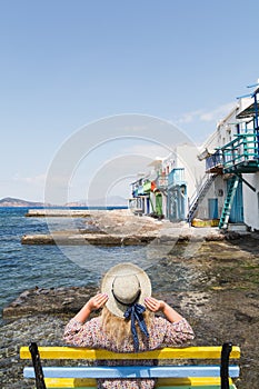 Young blonde girl sitting on a bench overlooking colourfull old houses in fishermen town of Klima on Milos island, Greece