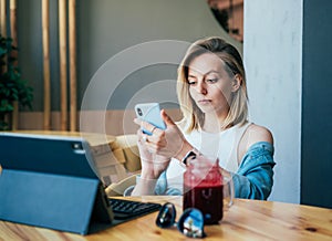 A young blonde girl sitting in a cafe looks at the social networks in the phone