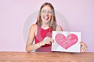 Young blonde girl sitting on the table holding heart draw sticking tongue out happy with funny expression