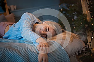 A young blonde girl smiles mysteriously and lies on the bed in a blue sweater.