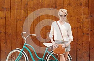 Young blonde girl is standing near the vintage green bicycle with brown vintage bag in orange sunglasses, warm, tonning