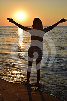 A young blonde girl stands on the beach and looks at the sun with her arms outstretched