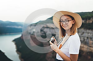 Young blonde girl in summer hat holds in hands on retro photo camera on background panorama horizin mountain and river landscape