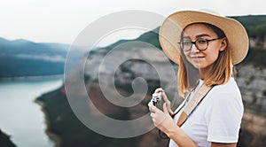 Young blonde girl in summer hat takes photo of  panorama horizin landscape on retro camera leisure during  trip on mountains