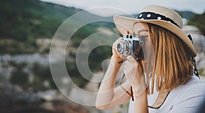 Young blonde girl in summer hat takes photo on retro camera on background panorama horizin mountain landscape, hipster tourist