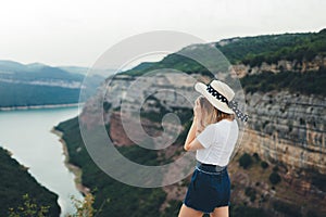Young blonde girl takes photo of  landscape on retro camera during  trip on mountains, hipster tourist in summer hat enjoys hobby
