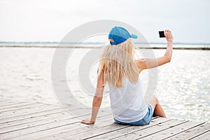 Young blonde girl taking selfie with phone on wooden pier