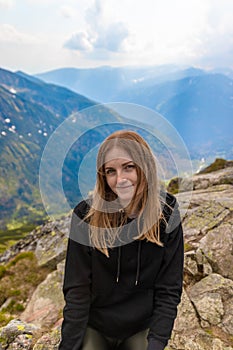 Young blonde girl traveler with a blue backpack raised and enjoying the green mountain scenery. Back view