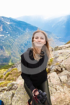 Young blonde girl traveler with a blue backpack raised and enjoying the green mountain scenery. Back view