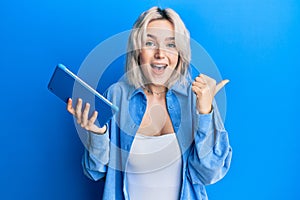 Young blonde girl using touchpad device pointing thumb up to the side smiling happy with open mouth