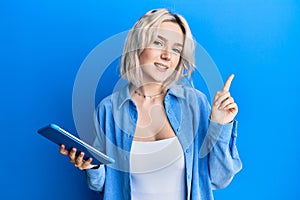 Young blonde girl using touchpad device smiling happy pointing with hand and finger to the side